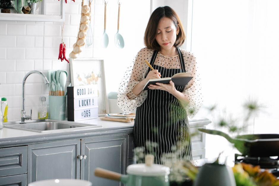 Asian woman in apron writing in notebook in a bright kitchen setting, surrounded by culinary items.